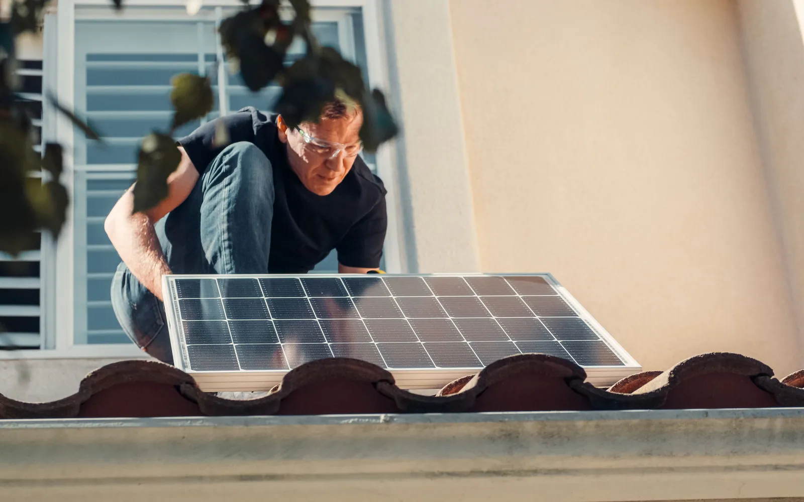 Installer in safety glasses setting a solar panel onto a clay tile roof beside a stucco wall