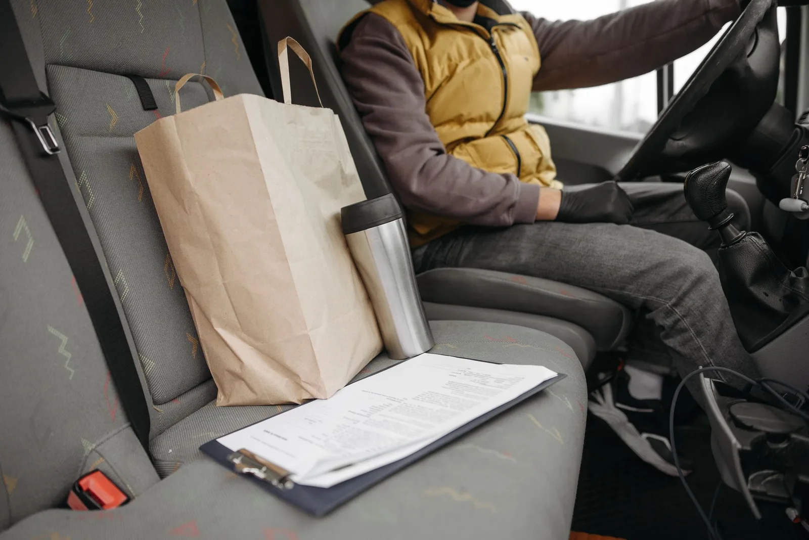A paper takeout bag and a travel mug on the passenger seat of a car while the driver holds the wheel
