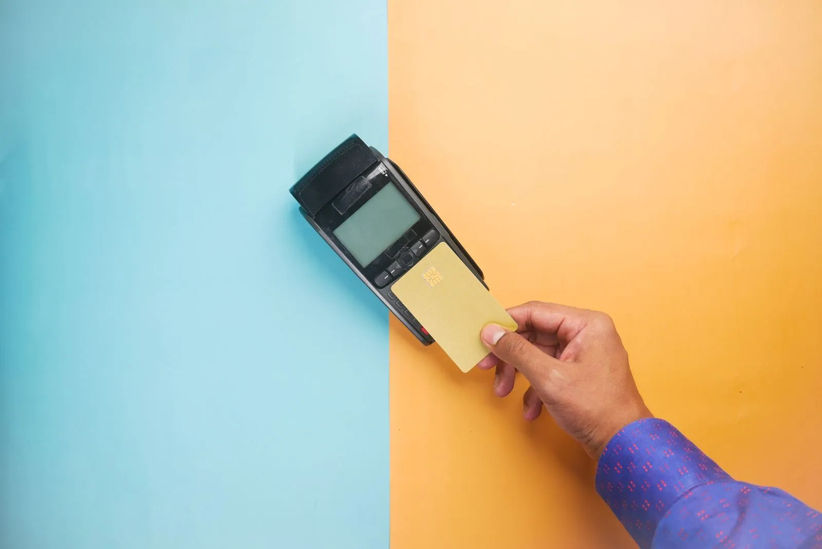 Hand holding a plain gold card to a payment terminal on a blue and orange background