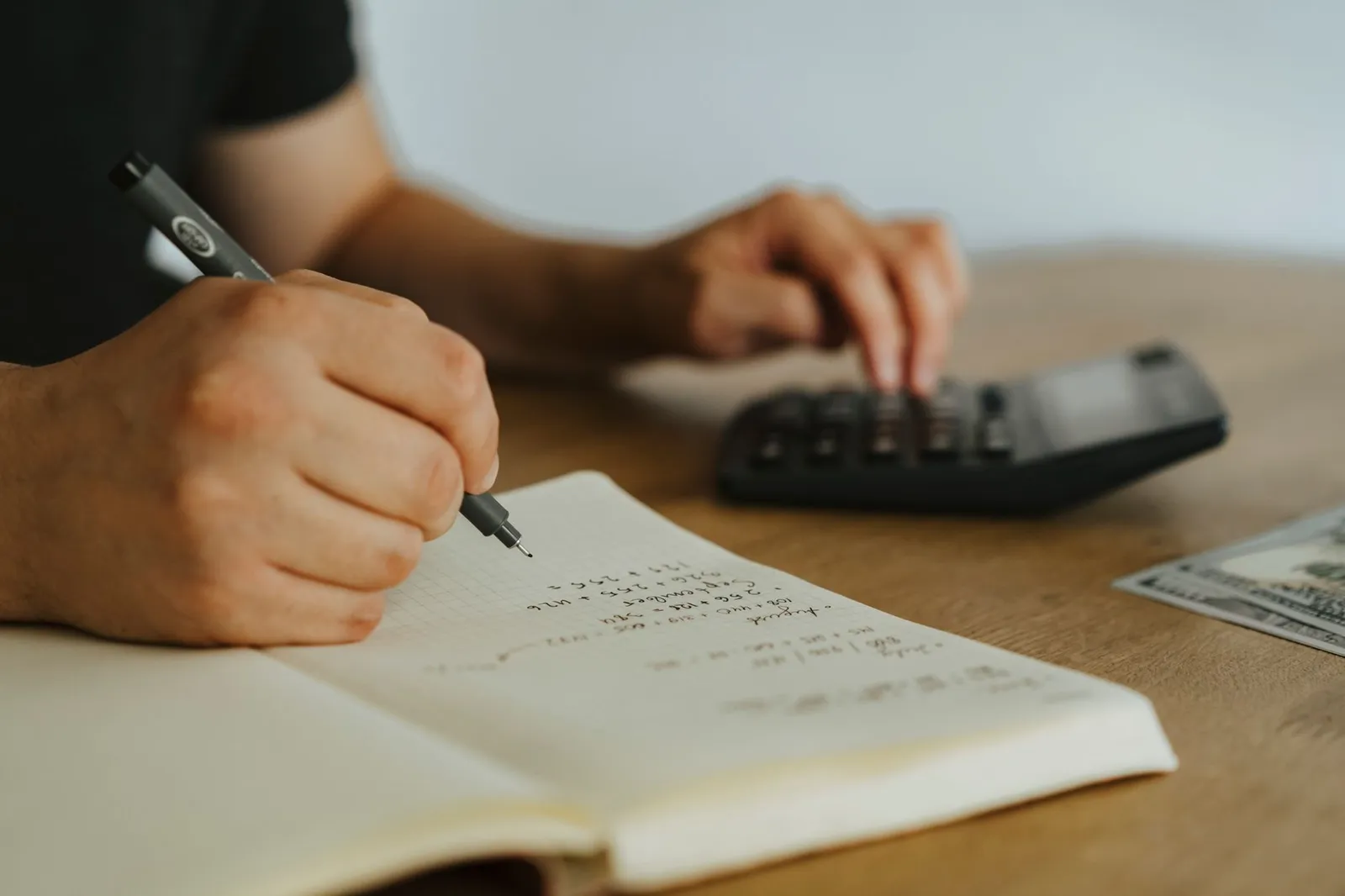 Person writing monthly calculations in a notebook beside a calculator on a wooden desk
