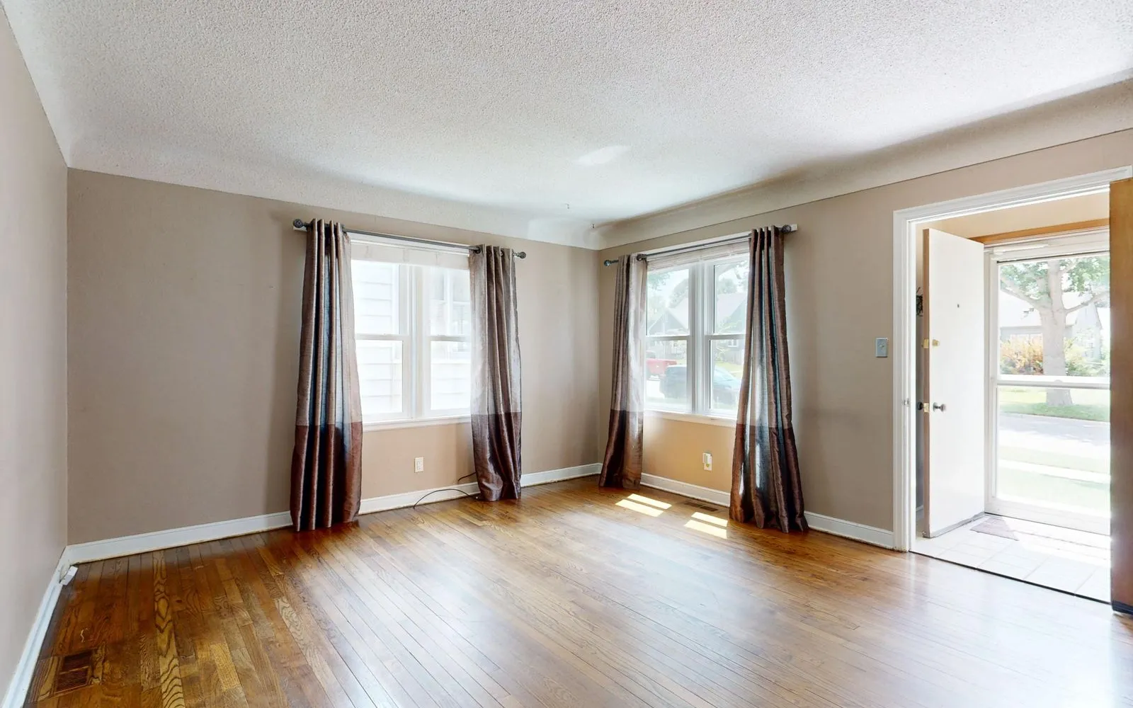 Empty living room with hardwood floors, curtained windows and the front door standing open