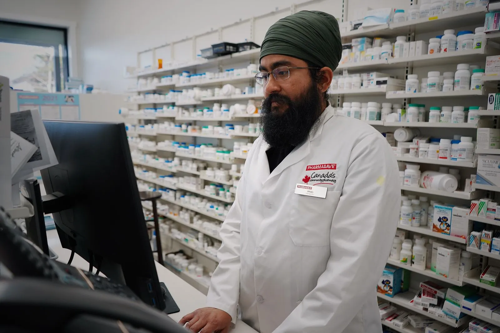A pharmacist working at a computer behind the pharmacy counter, with shelves of medicine in the background