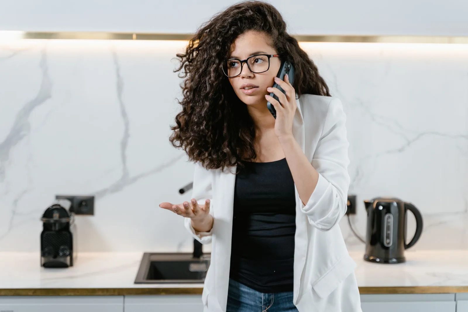 Woman in glasses on a phone call in a white kitchen, one hand raised mid-question