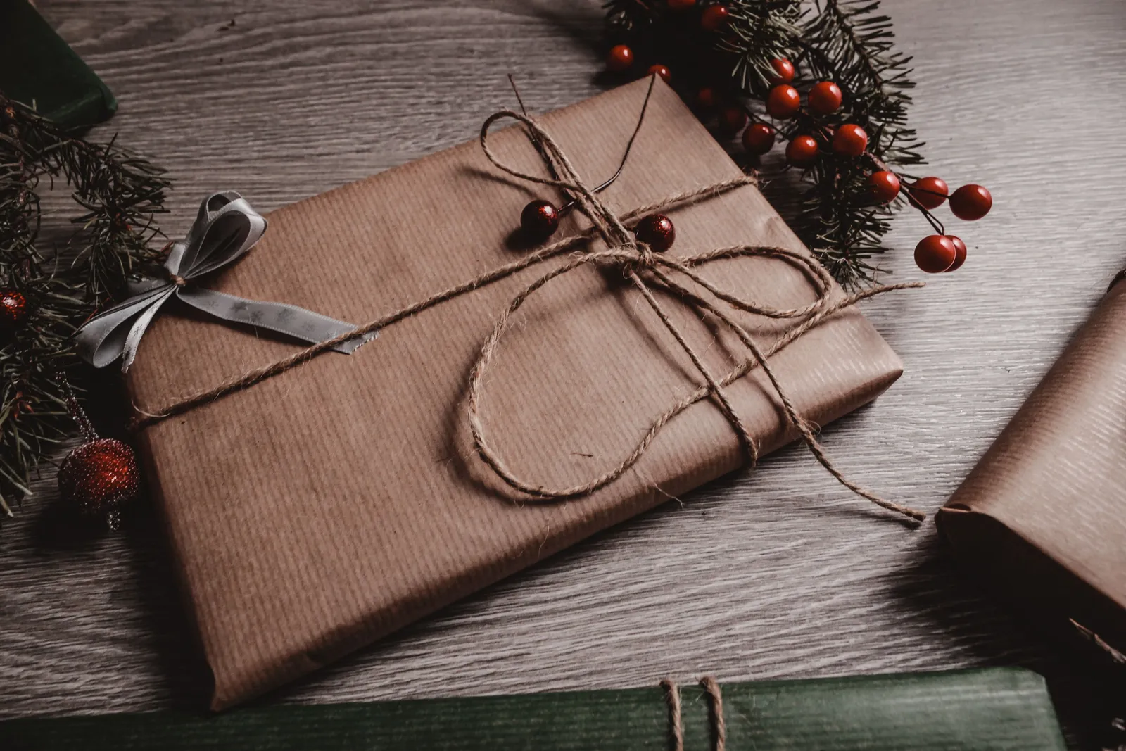 Gift wrapped in brown kraft paper and tied with twine on a wooden table, beside pine sprigs and red berries