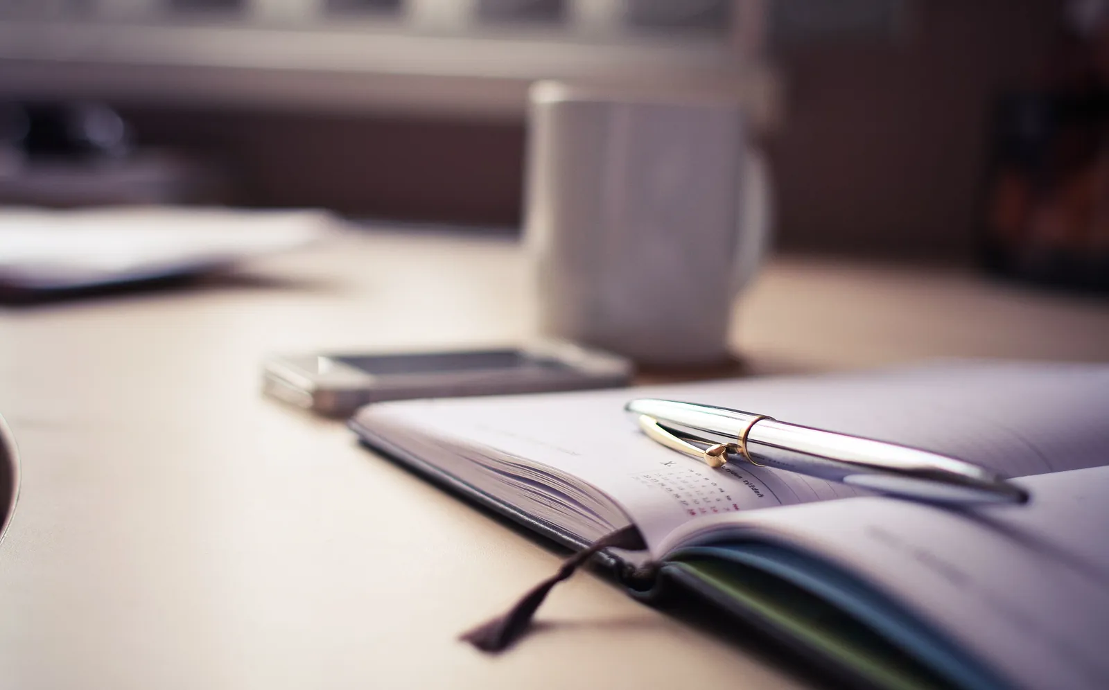 An open planner and pen on a desk beside a phone and a mug of coffee