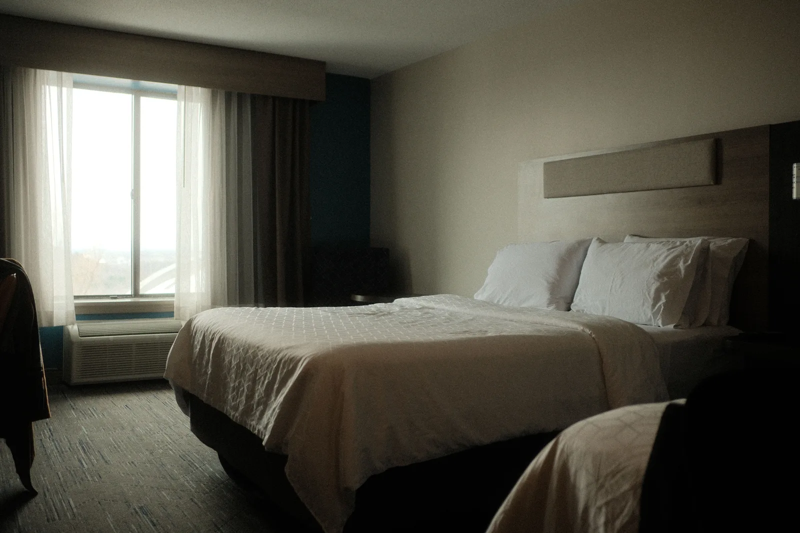 Dim hotel room with a made bed and sheer curtains over a window