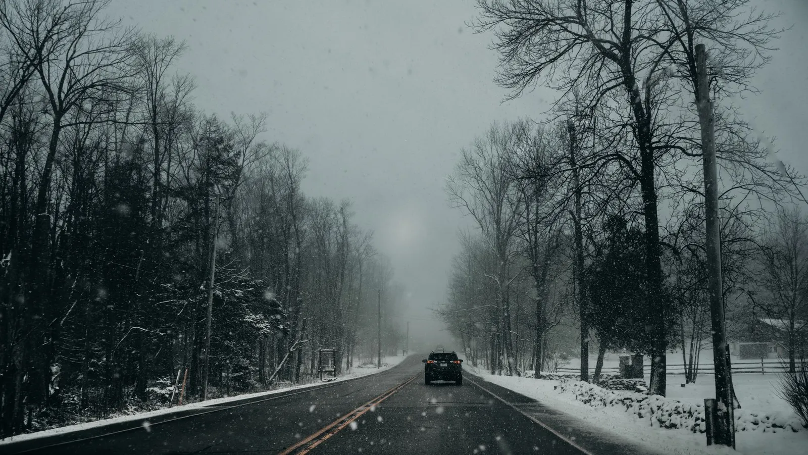 Car driving away on a two-lane road through bare trees in falling snow