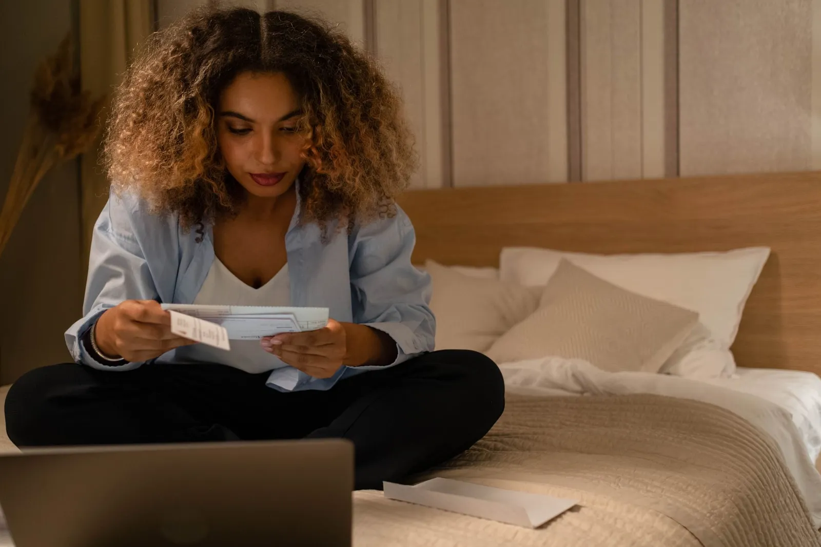 A woman sitting on a bed reading a mailed statement with a laptop open beside her