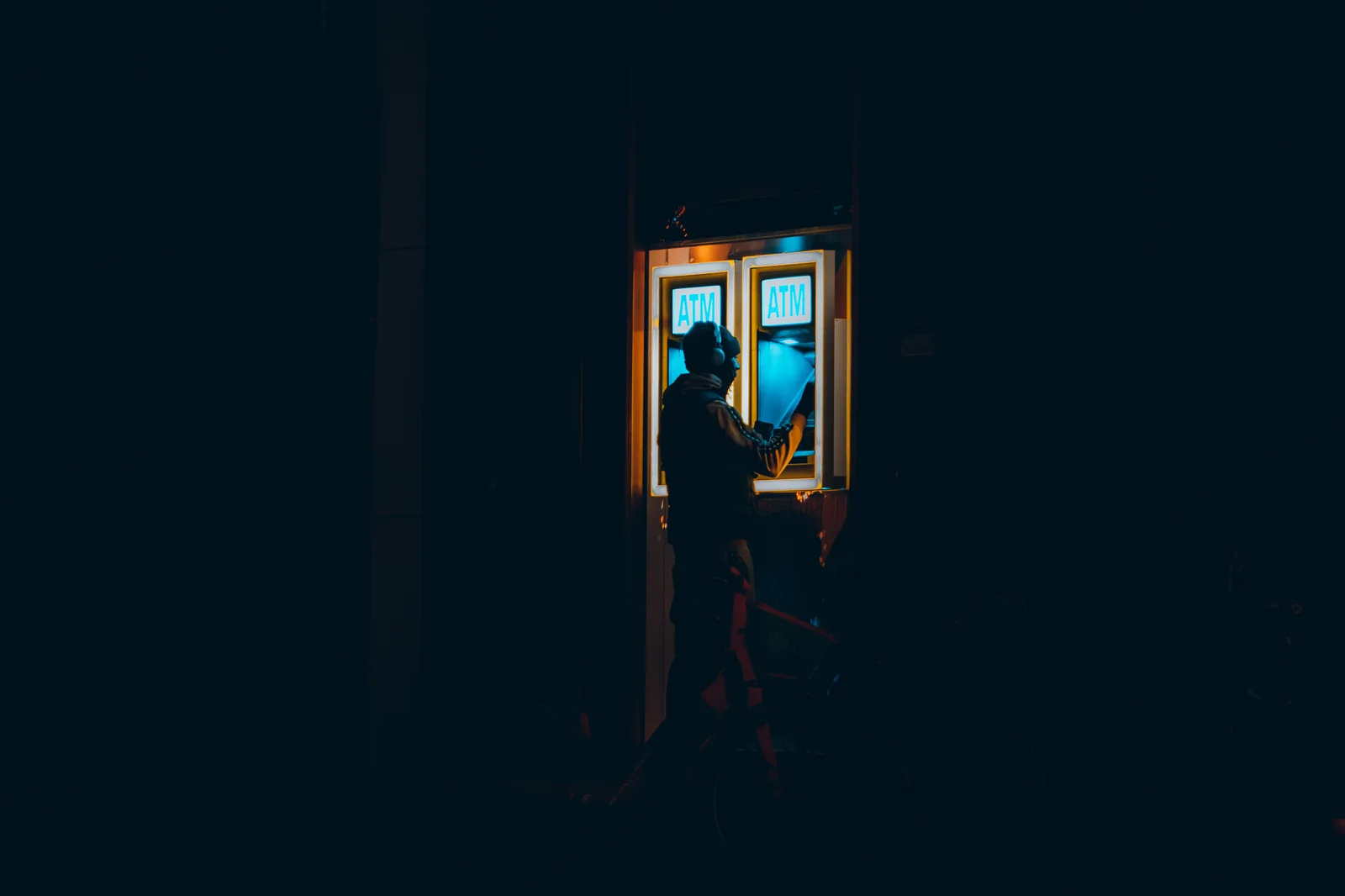A person standing at a lit cash machine on a dark city street at night