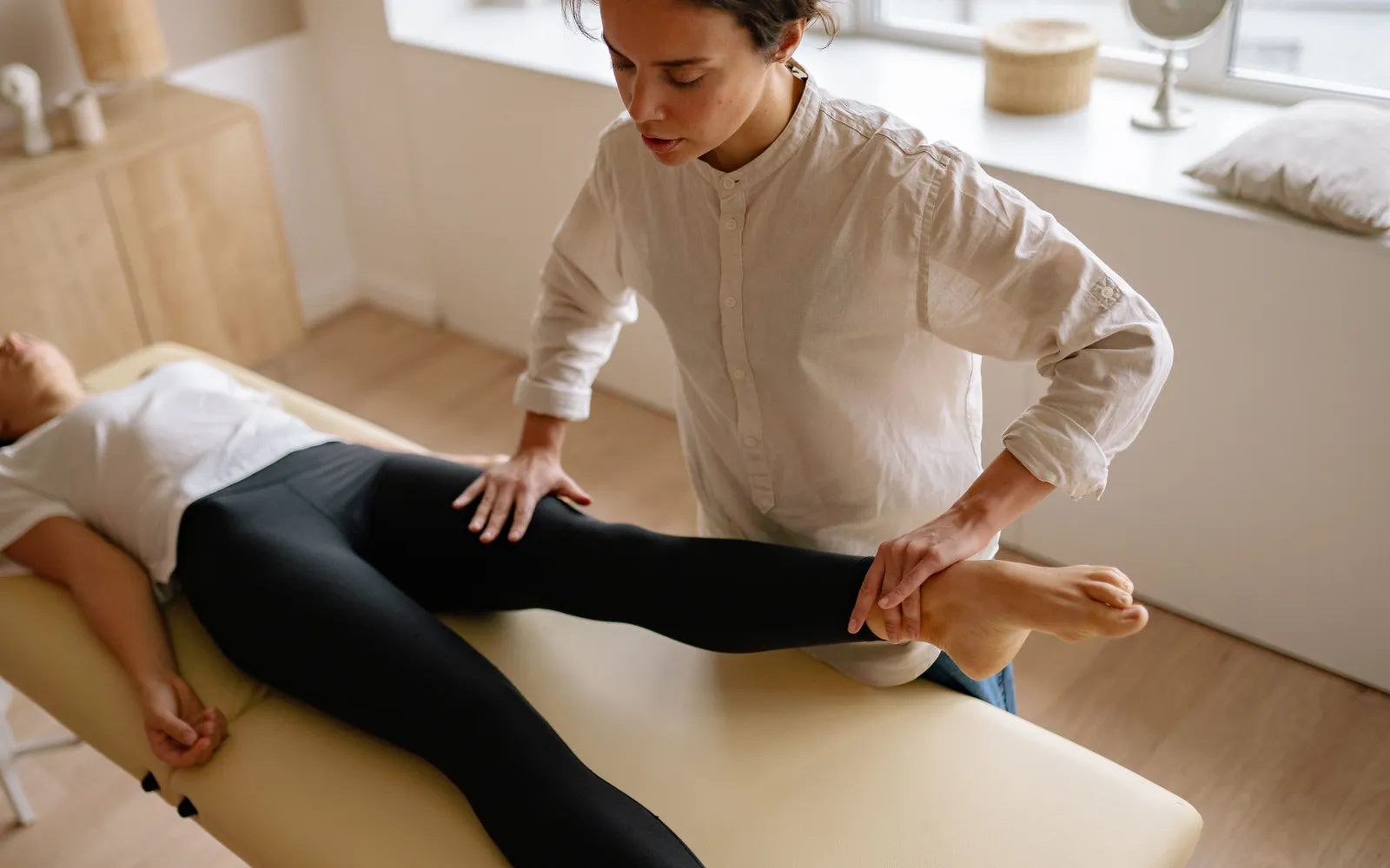 Physical therapist guiding a patient through a leg stretch on a treatment table in a bright clinic room