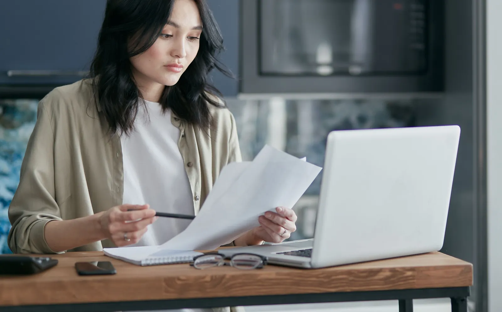 A woman reviewing paperwork at a home desk with a laptop open beside her