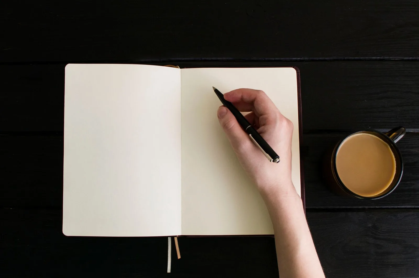 Hand holding a pen over a blank notebook beside a mug of coffee on a dark wooden table