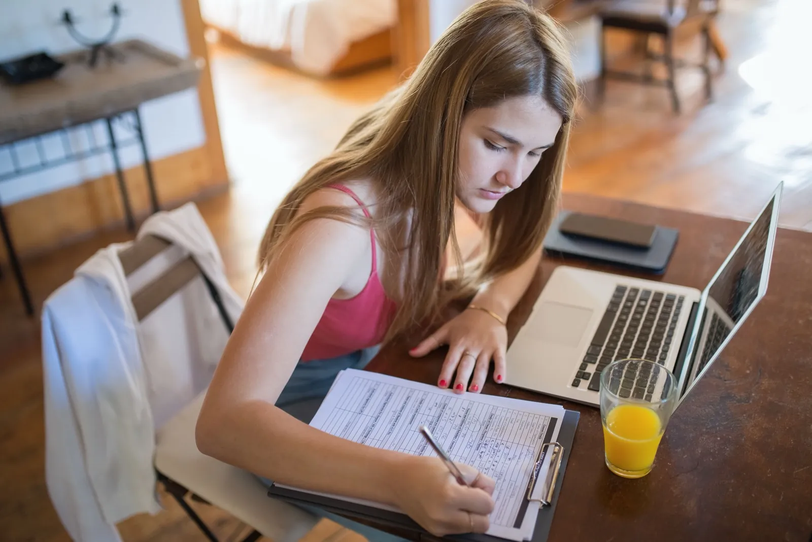 A young woman focused on a laptop at a home desk with a clipboard and notes