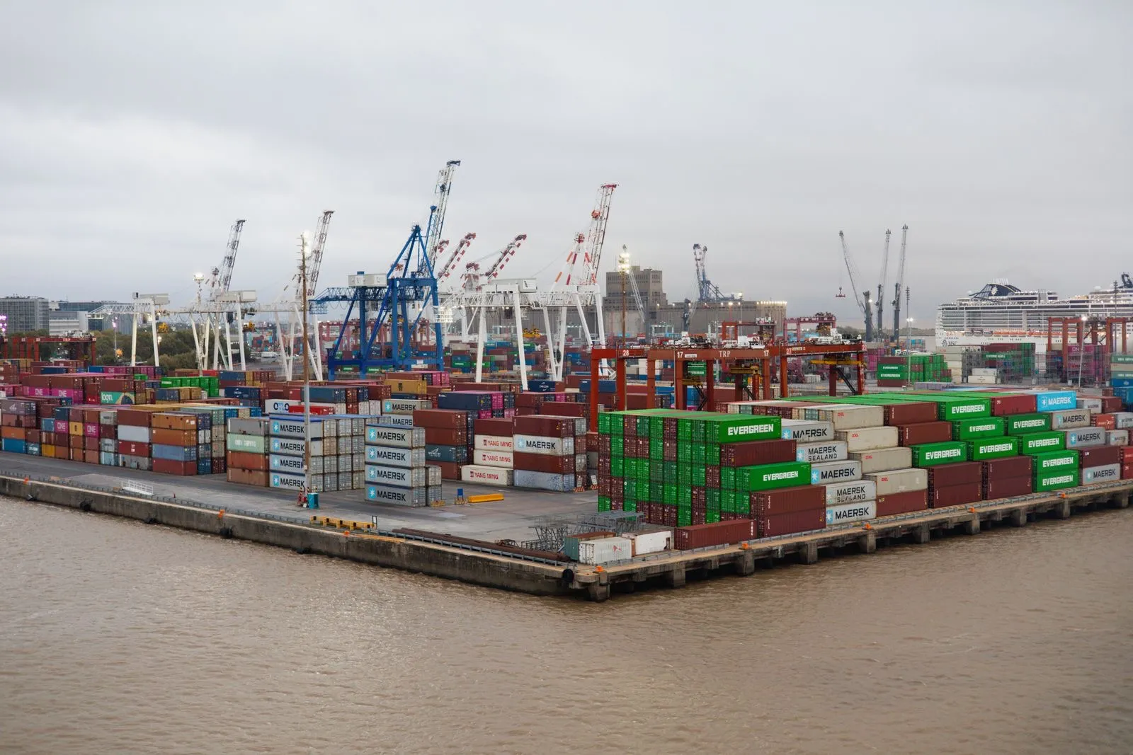 Stacked shipping containers and cranes at a busy port under an overcast sky