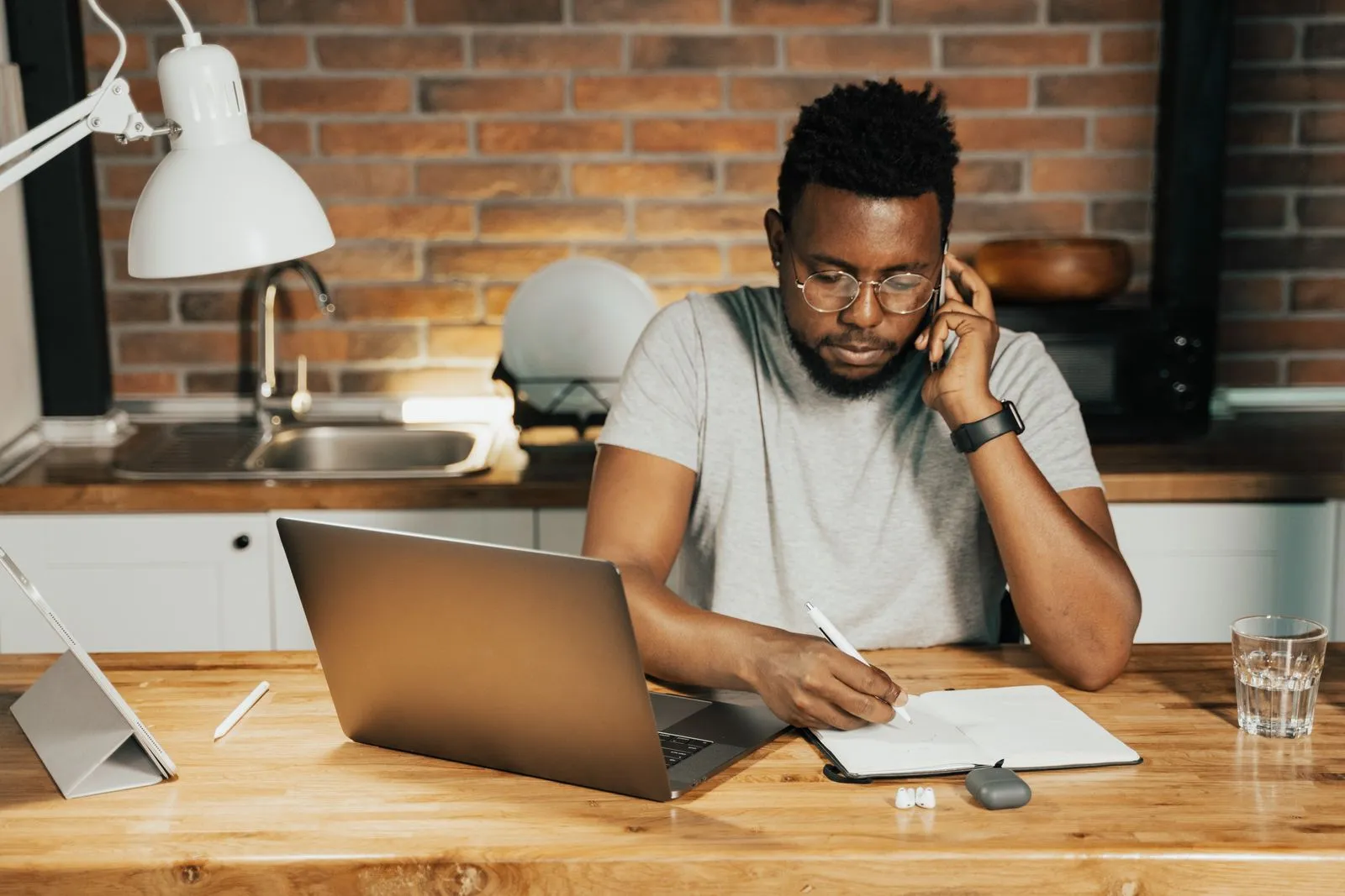 Man on a phone call taking notes at a kitchen table beside a laptop