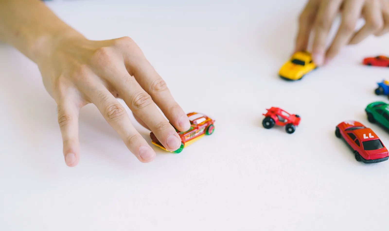 Hand resting on a red toy car with several other toy cars spread across a white table