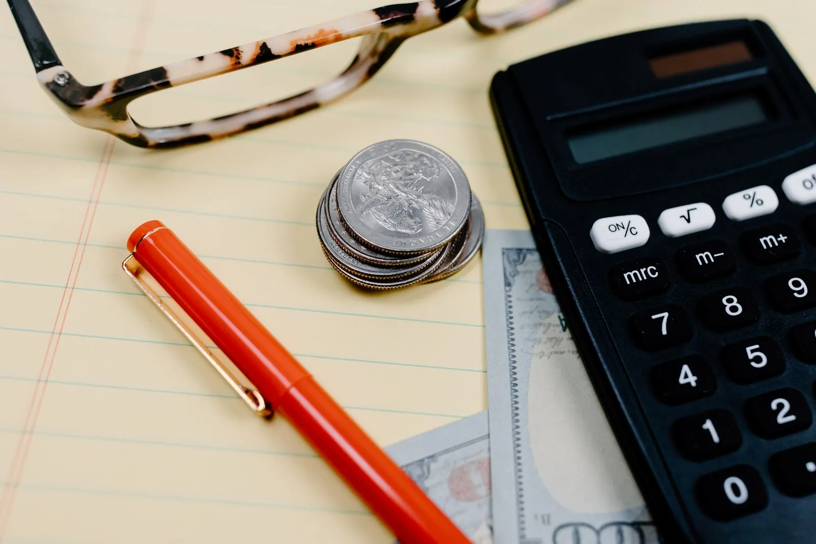 A calculator, coins, and reading glasses resting on a notepad on a desk