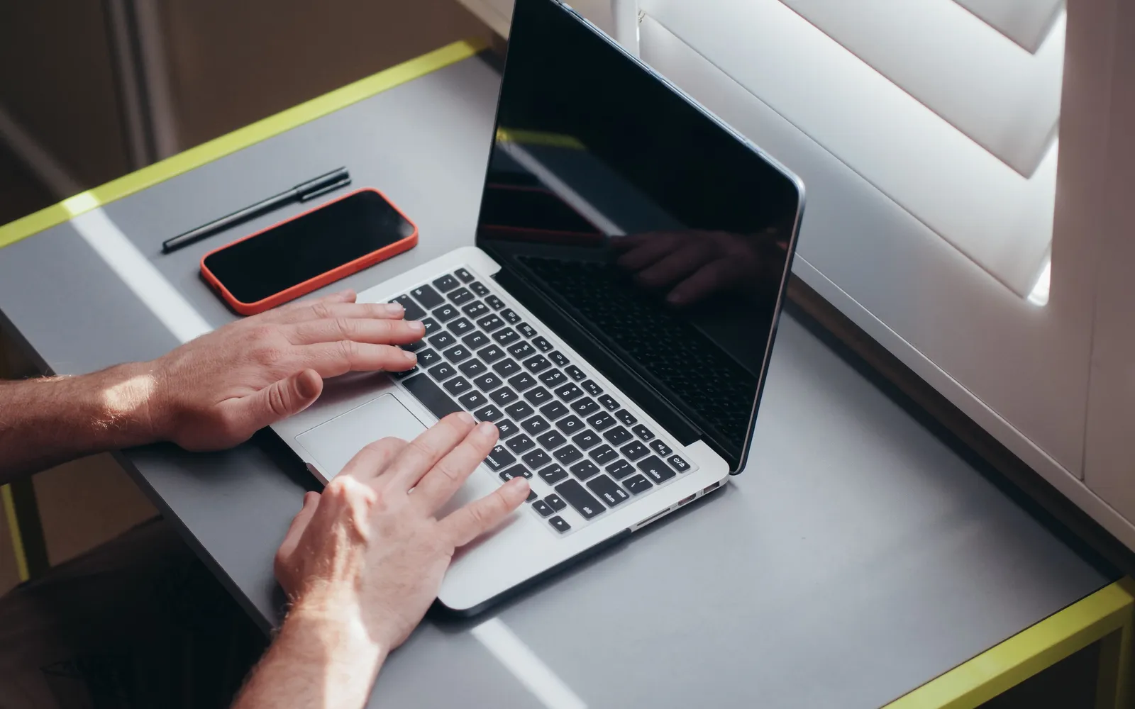 Hands typing on a laptop at a gray desk next to a phone and a pen by a window with shutters