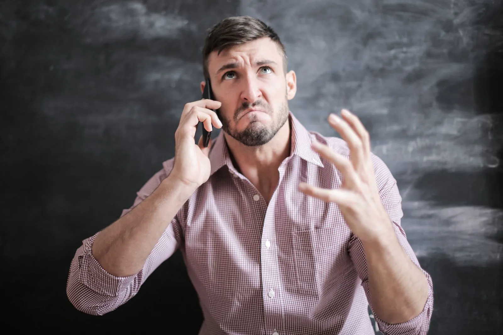 A man in a checked shirt on a phone call, eyes raised and one hand held up in exasperation, against a dark chalkboard