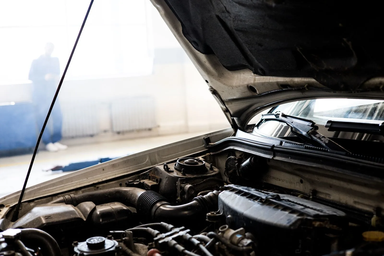 The open engine bay of a car in a bright garage, with a mechanic standing out of focus in the background
