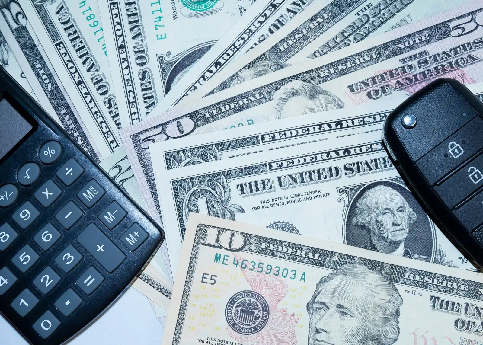 A fan of US dollar bills on a table with a black calculator and a car key fob resting on top