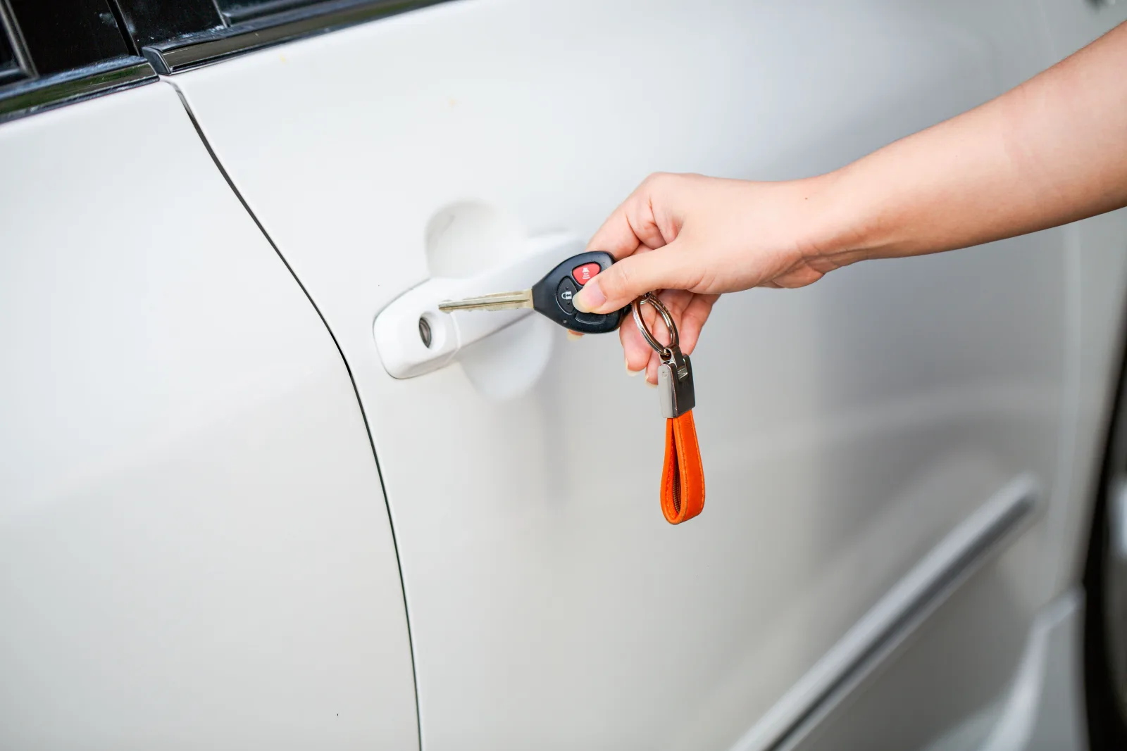 A hand holding a black key fob with an orange leather tag at the door handle of a white car