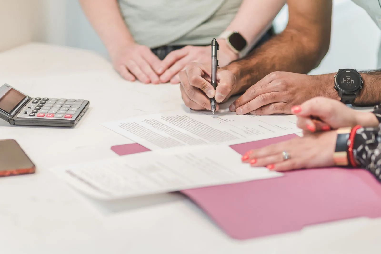 Two people at a white table signing a contract next to a calculator, a red folder, and a phone