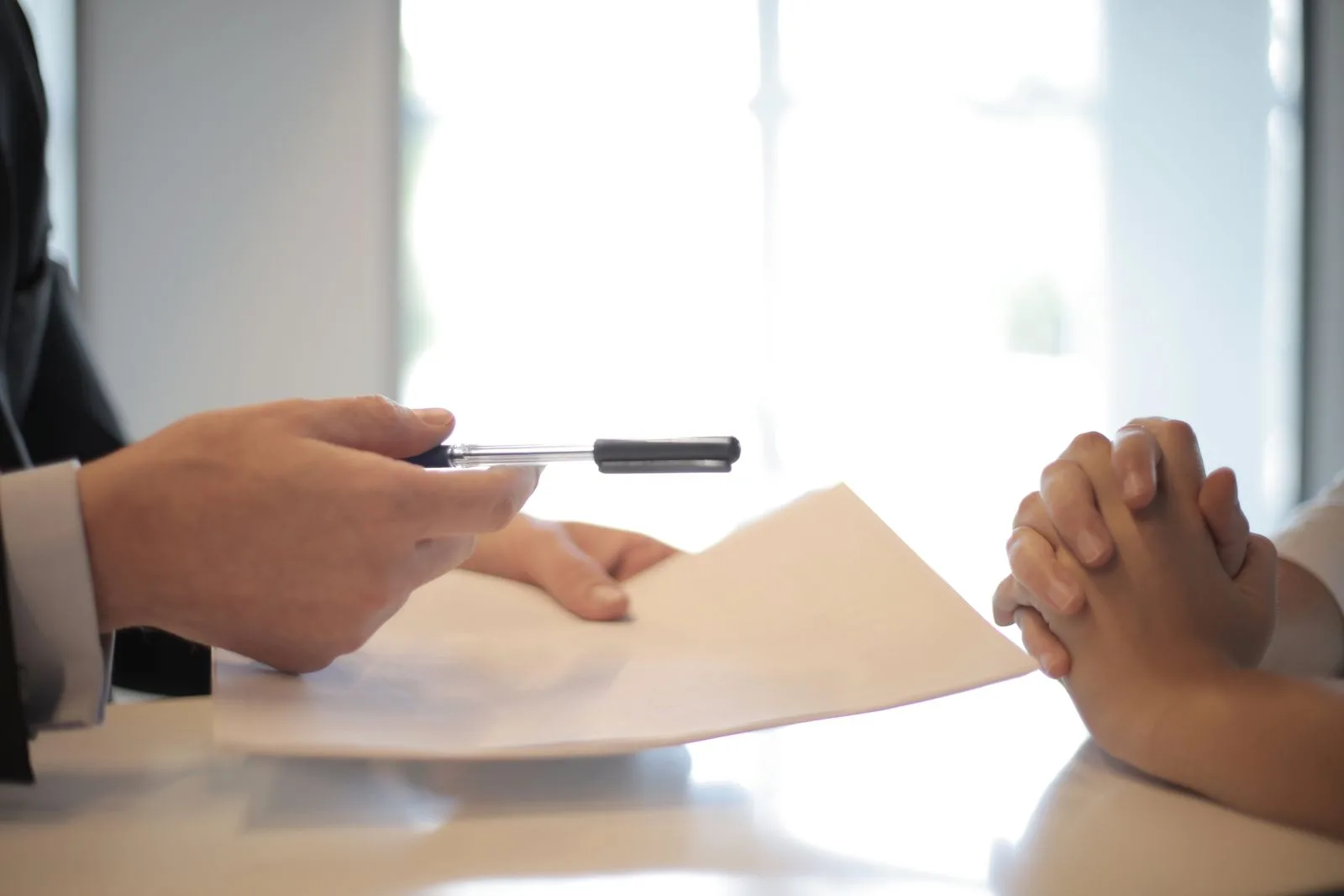 A salesman in a dark suit holding out a pen and a contract across a desk to a customer whose hands are clasped