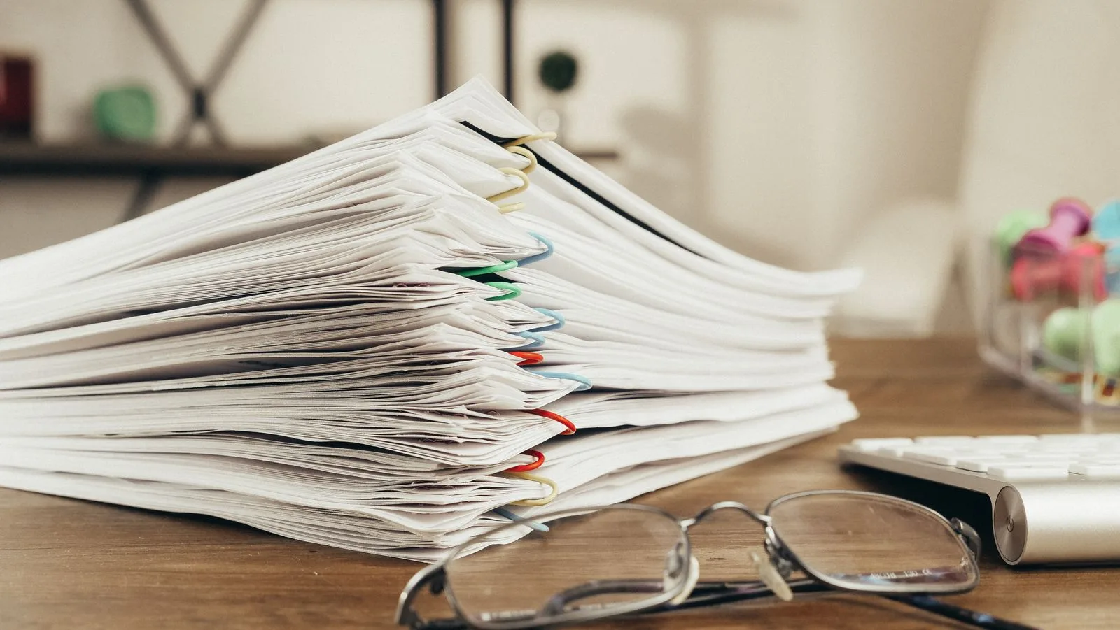 A thick stack of paper documents held together with colored paper clips, with a pair of reading glasses in front