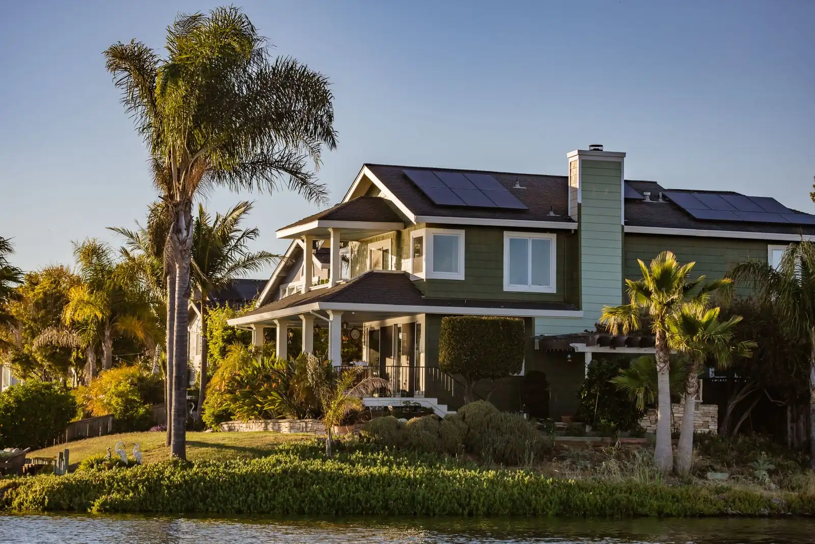 A two-story clapboard house with solar panels across its roof, seen from across the water at dusk