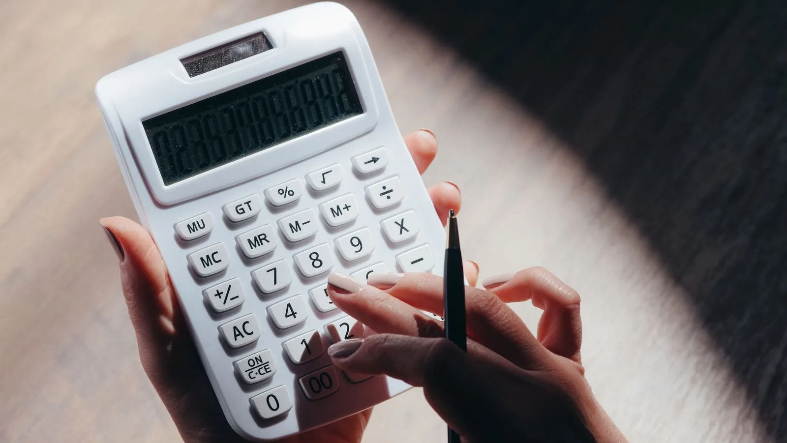 A pair of hands holding a plain white calculator and a pen above a wooden table